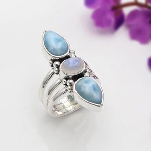 May include: A silver ring featuring three vertically aligned stones. The top and bottom stones are teardrop-shaped, opaque, light blue Larimar. The center stone is a round, translucent, iridescent moonstone. The band consists of three silver wires.