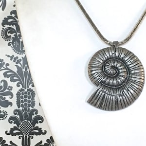 May include: A silver-toned pendant necklace featuring a large, spiral-shaped pendant. The pendant has a textured surface with detailed lines. The necklace is displayed against a white background with a patterned fabric on the left side.
