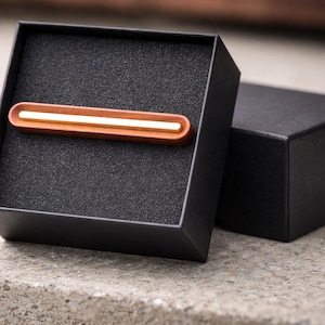 May include: A wooden tie bar with a gold-coloured accent and a white stripe, presented in an open black box with a foam insert. A second black box is visible in the background, resting on a concrete surface.