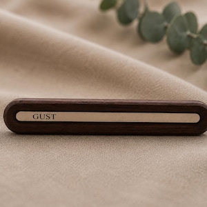 May include: A dark brown wooden pen holder with a rectangular, metallic accent. The word "GUST" is engraved on the metallic strip. The pen holder is set against a neutral-toned fabric background, with green leaves in the upper right corner.