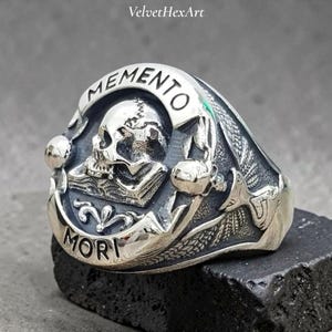 May include: A silver ring with a skull design. The ring features the words "MEMENTO MORI" and a detailed skull image. The ring is set on a dark surface. The ring is likely made of sterling silver.