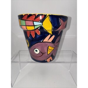 May include: A ceramic flower pot with a dark blue background featuring hand-painted colorful fish, a yellow shape, and a red patterned element. The rim is yellow, and the design includes a large purple fish with a white eye and yellow fin.