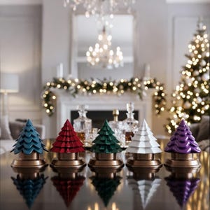 May include: Five colorful Christmas tree decorations in teal, red, green, white, and purple. Each tree sits on a gold-colored base. The trees are arranged on a reflective surface, with a Christmas tree and fireplace in the background.