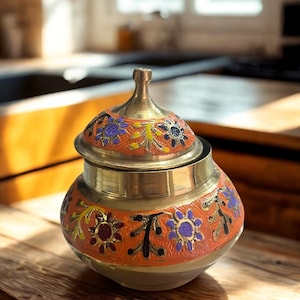 May include: A decorative, lidded jar with a brass finish and intricate floral designs. The jar features a vibrant orange band with purple, yellow, and maroon flower patterns. The jar sits on a wooden surface.