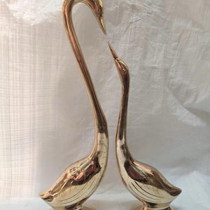 May include: Two brass swan figurines. One swan has a curved neck, the other has a straight neck. Both swans are on a round base with the word "SMIT" on the base. The swans are gold in color and are displayed on a white background.