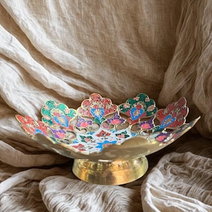 May include: A decorative brass bowl with a floral design. The bowl has a wide, scalloped rim and a round base. The floral design features red, blue, green, and pink colors. The bowl is set against a neutral fabric background.