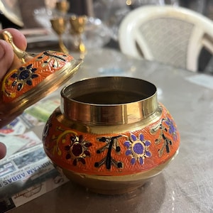 May include: A small, round, orange and gold decorative jar with a lid. The jar features floral designs in purple, blue, and red. The lid is open, revealing the inside of the jar. The jar is made of metal.