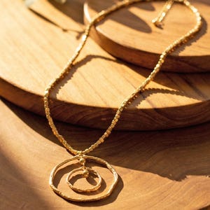 Op de afbeelding: Een goudkleurige ketting met een snoer van kleine, vierkante kraaltjes. De hanger bestaat uit twee concentrische, onregelmatige cirkelvormige ringen met een getextureerde, gehamerde afwerking, verbonden door een klein staafje met twee bolletjes.