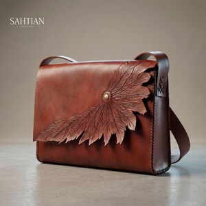 May include: A brown leather messenger bag with a carved feather design on the flap. The bag has a shoulder strap and a brass button closure. The brand name "SAHTIAN ISTANBUL" is printed on the top left.