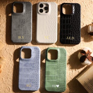 May include: Five rectangular phone cases with a crocodile-embossed texture in grey, white, black, light blue, and green. Each case features a camera cutout and gold-coloured personalised initials or text, including B.Y, VC, J.E.D, AG, and HI with a heart.