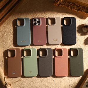 Puede incluir: Una colección de nueve fundas para teléfono texturizadas en varios colores, incluyendo azul, malva, beige, negro, marrón, verde claro, gris oscuro, salmón y verde oliva. Cada funda tiene un recorte para la cámara y algunas incluyen iniciales.