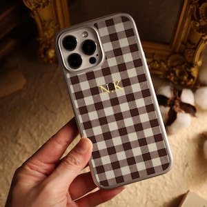 May include: A smartphone case with a brown and white gingham design. The case has a silver frame and gold initials "N.K." on the back. The case is designed for a mobile phone.