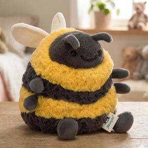 May include: A plush bumblebee toy with a round body, featuring alternating bands of yellow and dark gray. It has small, gray antennae, wings, and legs. A small tag with the brand name is attached to the toy.