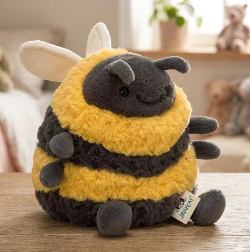 May include: A plush bumblebee toy with a round body, featuring alternating bands of yellow and dark gray. It has small, gray antennae, wings, and legs. A small tag with the brand name is attached to the toy.