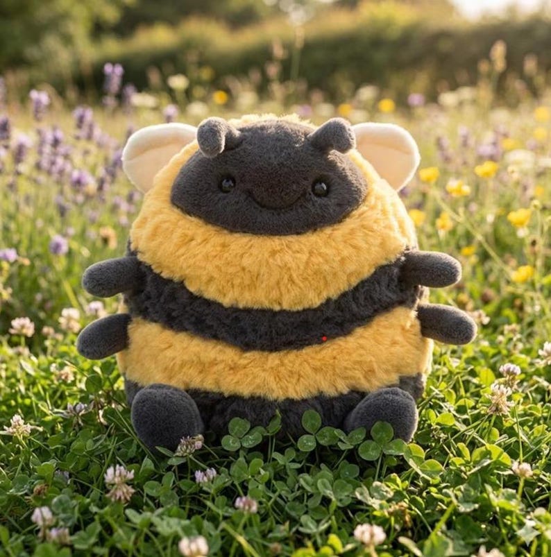 May include: A plush bee toy with a round body, featuring yellow and black stripes. The bee has small, gray arms and legs, with a smiling face and small antennae. The toy is set in a field of green grass and wildflowers.