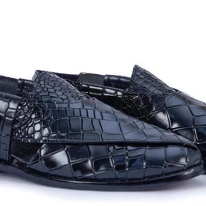 Pode incluir: Um par de sapatos slingback em azul-marinho escuro com uma textura brilhante de relevo de crocodilo. Os sapatos têm bico fino, salto baixo e uma tira lateral com fecho de fivela metálica, apresentando um design estruturado.