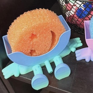 May include: Two whimsical sponge holders in the shape of smiling characters. One holds an orange sponge, the other a blue sponge. The holders are light blue and pink, with matching feet and hands. A fun and functional kitchen accessory.