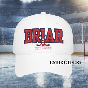 May include: White baseball cap with the word "BRIAR" in red block letters with a blue outline, and crossed hockey sticks. The word "UNIVERSITY" is below the hockey sticks. The word "EMBROIDERY" is below the cap.