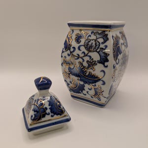 Peut inclure: Un pot en céramique blanche à base carrée avec son couvercle assorti, orné de motifs floraux bleus et de bordures dorées. Le pot présente un corps fuselé et un couvercle carré avec un petit bouton, tous deux décorés de motifs bleus.
