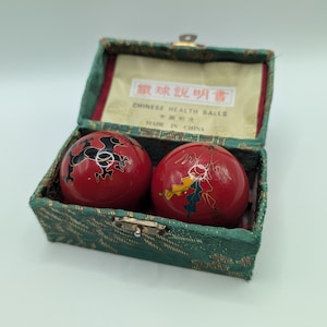Peut inclure: Deux boules de santé chinoises sphériques rouges avec des motifs graphiques noirs et multicolores, présentées dans une boîte rectangulaire recouverte de tissu vert. L'intérieur du couvercle porte une étiquette avec le texte Chinese Health Balls.