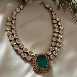 Puede incluir: Un collar dorado con dos hileras de piedras claras y redondeadas y una gran piedra central rectangular de color verde esmeralda. El collar tiene una estética vintage y se presenta sobre una superficie blanca. El texto "TRUSTWITHUSA" es visible.