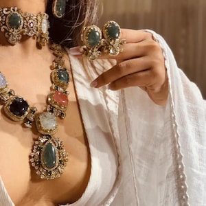 Op de afbeelding: Een sierlijke sieradenset met een ketting, oorbellen en ring. De ketting en oorbellen hebben grote, kleurrijke edelstenen in goudkleurig metaal. De ring heeft twee groene edelstenen. De tekst "TRUSTWITHUSA" is zichtbaar.