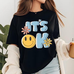 May include: Black t-shirt featuring the phrase "IT'S OK" in light blue bubble letters, a yellow smiley face, and floral accents. A casual, comfortable style.