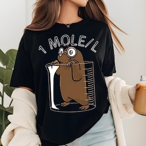May include: Black t-shirt featuring a cartoon mole wearing glasses inside a beaker. The text "1 MOLE/L" is printed above the mole. The mole is brown with white eyes and a white belly. The beaker has measurement lines.