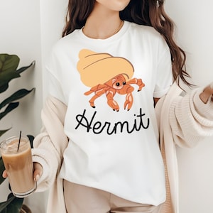 Può includere: T-shirt bianca con un disegno di paguro dei cartoni animati. Il granchio ha un corpo arancione e un guscio marrone. La parola "Hermit" è scritta in corsivo nero sotto. Un capo casual per tutti i giorni.
