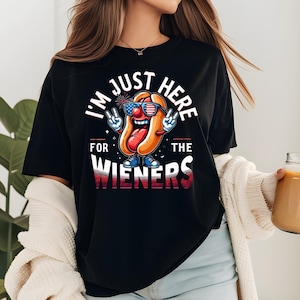 Può includere: T-shirt nera con la scritta "I'M JUST HERE FOR THE WIENERS" e un disegno di un hot dog a cartoni animati. L'hot dog indossa occhiali da sole e scarpe, con la lingua di fuori e segni di pace. Il design utilizza i colori rosso, bianco e blu.