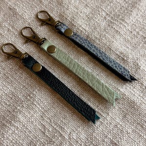 Op de afbeelding: Drie lange, smalle sleutelhangers met getextureerde oppervlakken in zwart, lichtgroen en een donkergrijs slangenleerpatroon. Elke hanger is voorzien van een messingkleurige metalen karabijnhaak en een bijpassende drukknoop bij het bevestigingspunt.