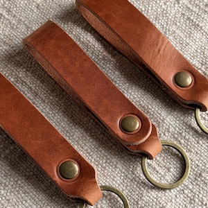 May include: Three brown leather key fobs arranged diagonally. Each strap is folded and secured with a circular brass-toned metal snap fastener. A matching brass-toned metal split ring is attached to the bottom of each leather loop.