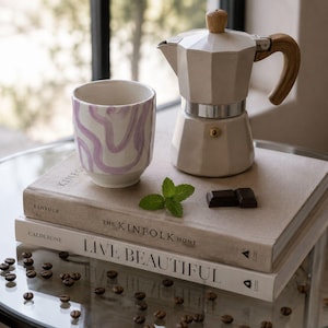 Peut inclure: Nature morte sur le thème du café. Une cafetière italienne beige clair avec une poignée et un bouton en bois est posée à côté d'une tasse blanche avec un motif tourbillonnant violet. Des livres avec les titres "KINFOLK" et "LIVE BEAUTIFUL" sont empilés.