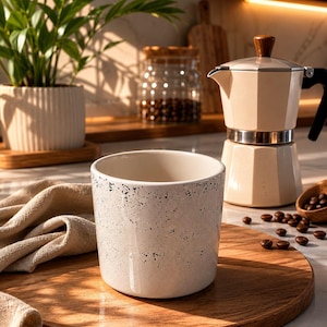 Peut inclure: Une tasse en céramique blanche mouchetée de noir est posée sur une planche à découper en bois. Une cafetière beige et des grains de café sont en arrière-plan. Une petite plante en pot est également visible.