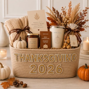 May include: A beige rope basket with the text THANKSGIVING 2026 in gold rhinestones, filled with a beige knit blanket, a candle, a chocolate bar, a tumbler, a bag of chocolates, a card, and dried autumn foliage with a small white pumpkin.