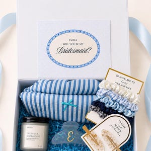 May include: A white gift box filled with bridesmaid proposal items. Includes a card that says "EMMA, WILL YOU BE MY Bridesmaid?", a striped sleep mask, scrunchies, hair clips, a hair claw clip, and a candle.