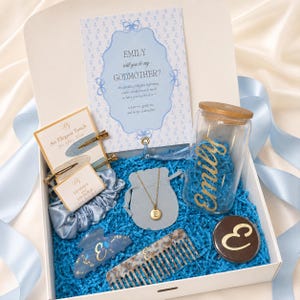 May include: A gift box containing a card asking, "Emily will you be my Godmother?", a glass tumbler with a bamboo lid and the name "Emily" in gold, a gold necklace with an "E" pendant, a blue scrunchie, a hair clip, a comb, and a compact mirror.