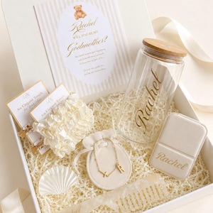 May include: A gift box containing a card with a teddy bear graphic and the text Will You Be My Godmother, a glass tumbler with a bamboo lid labeled Rachel, a cream scrunchie, a gold initial bracelet in a pouch, a compact mirror, and a comb.