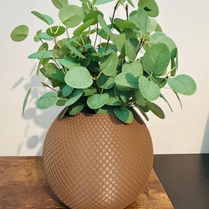 May include: A brown, spherical vase with a textured, cross-hatched pattern, filled with green, round-leafed faux eucalyptus. The vase sits on a wooden surface, creating a natural, decorative accent for home decor.