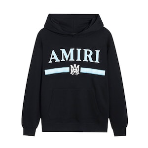 May include: Black hooded sweatshirt with the word "AMIRI" in white and light blue lettering across the chest. Below the word "AMIRI" are two light blue stripes and a white emblem. The sweatshirt has a front pocket.