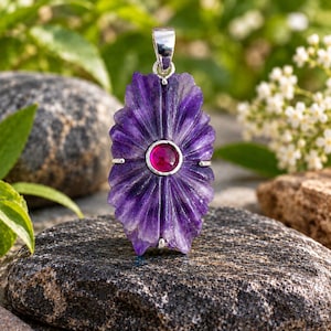 May include: A pendant featuring a carved, deep purple amethyst stone with a radial, flower-like pattern. A small, round, vibrant pink gemstone is set in the centre of the amethyst. The piece includes a polished silver-toned metal bail at the top.