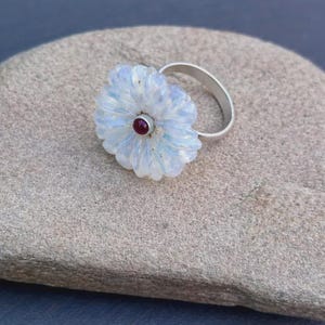 May include: A silver ring featuring a translucent, pale blue, flower-shaped stone with a small, round, deep red gemstone set in the center. The flower petals have a carved, textured appearance, and the band is a simple, polished silver metal.