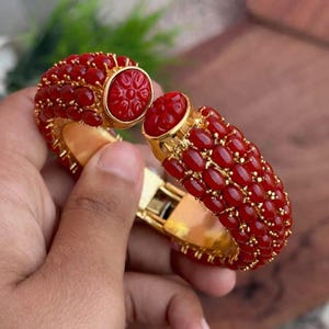 Puede incluir: Un brazalete rígido articulado de tono dorado con tres filas de piedras rojas ovaladas en cabujón. Los extremos del brazalete están rematados con piedras rojas circulares más grandes, talladas con un patrón floral y detalles dorados.