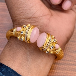 May include: A gold cuff bracelet featuring two large, oval, light pink cabochon stones at the open ends. The metal surface has a textured, ribbed design and is accented with smaller, round, clear white stones set in intricate, ornate gold metalwork.