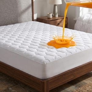 May include: A white quilted mattress protector on a wooden bed frame. Orange liquid is being poured onto the mattress, creating a splash. The bed is in a bedroom setting with a nightstand and lamp visible in the background.