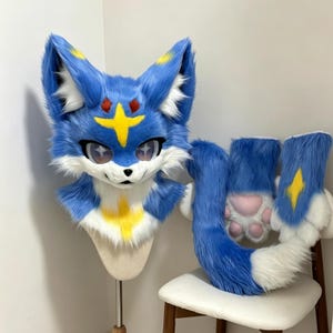 May include: A blue and white faux fur animal head mask featuring large pointed ears with yellow accents, a yellow star-shaped forehead marking, and star-shaped pupils. The set includes matching blue and white paw gloves with pink paw pads.