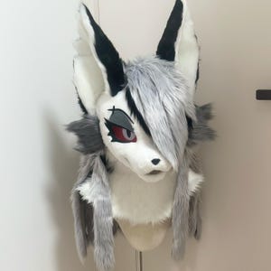 May include: A faux fur animal head mask featuring long pointed ears with black and white fur. The mask has grey and white fur, red eyes with dark grey eyeshadow, a black nose, and a side-swept fringe of grey hair covering one eye.