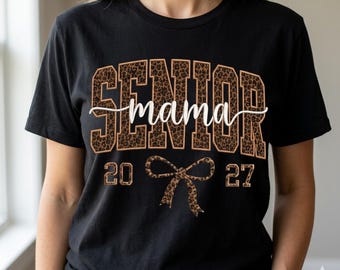 May include: A black short-sleeved t-shirt featuring the word SENIOR in large block letters with a brown and black leopard print pattern. The word mama is written in white cursive script across the centre, with 2027 and a leopard print bow.