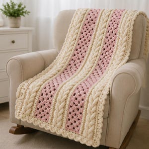 May include: A crochet blanket featuring alternating vertical stripes of cream-coloured cable knit patterns and light pink textured granny stitch sections. The blanket has a scalloped edge border and is draped over a neutral-coloured rocking chair.