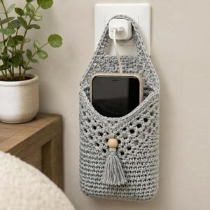 Könnte beinhalten: Ein hellgrauer, gehäkelter Handyhalter mit Lochmuster, einer einzelnen Holzperle und einer passenden grauen Quaste. Der Halter hängt an einer weißen Wandsteckdose und enthält ein schwarzes Smartphone, das an ein weißes Ladegerät angeschlossen ist.